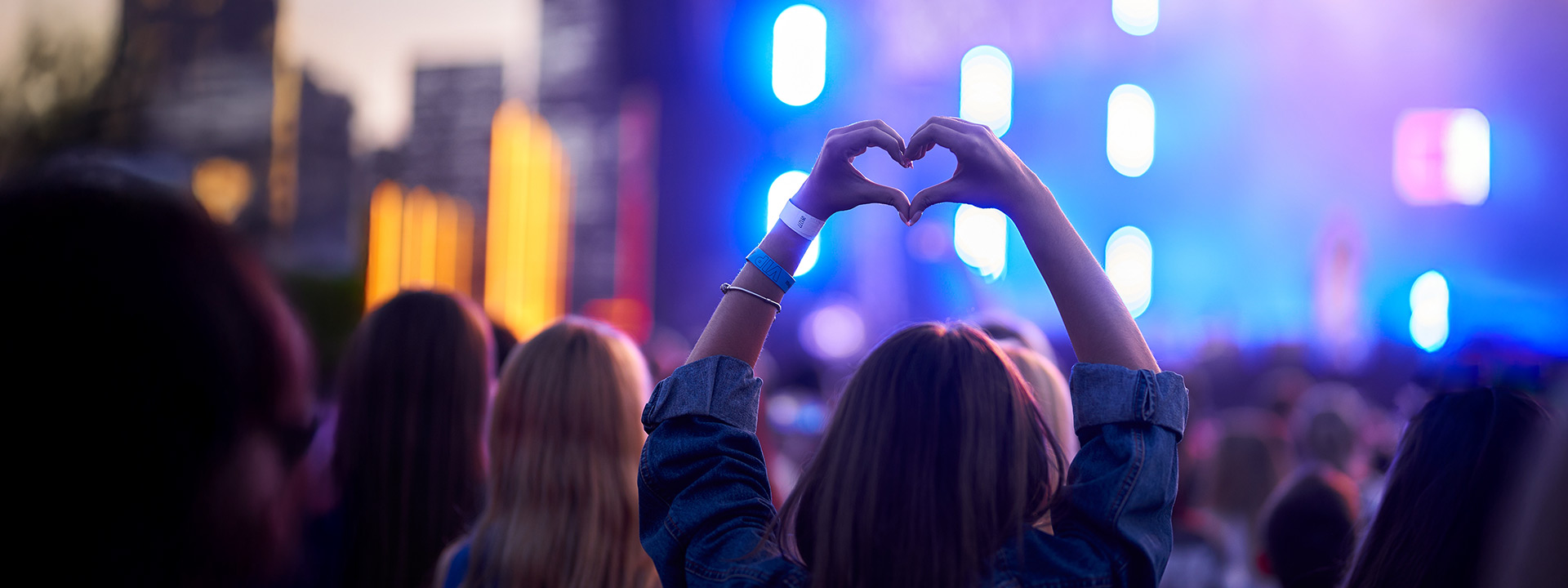 A person in a crowd at an outdoor concert forms a heart shape with their hands as blue and purple stage lights illuminate the background.