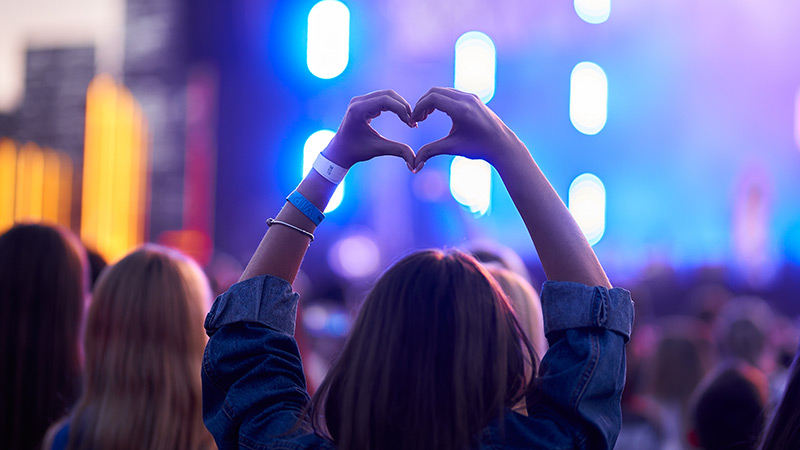 A person in a crowd at an outdoor concert forms a heart shape with their hands as blue and purple stage lights illuminate the background.