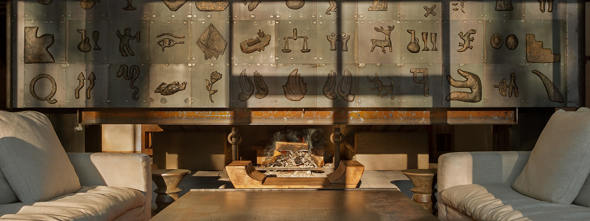 A cozy living room with two beige armchairs and a coffee table facing a modern fireplace. The wall behind features metal art with various abstract shapes and symbols.