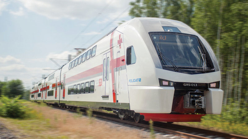 A sleek, modern double-decker passenger train with white and red stripes travels on a track through a scenic area with trees and clear skies.