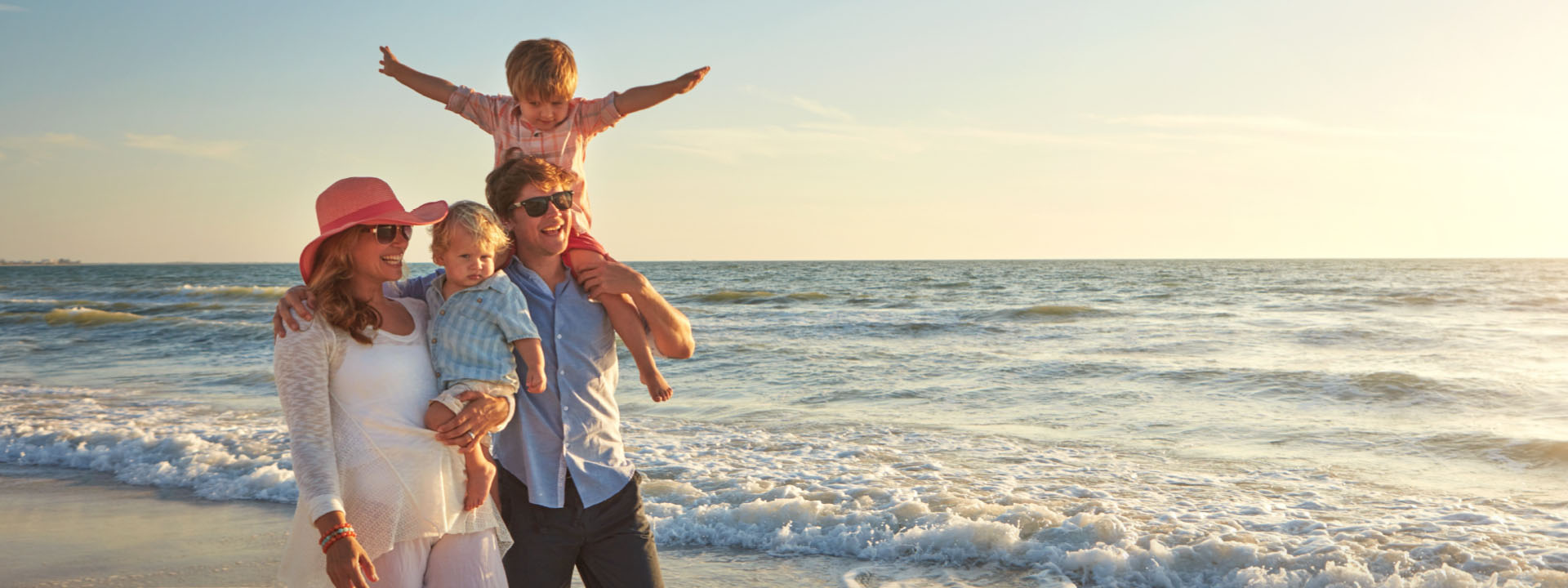 A family of four stands on the beach at sunset; two adults hold a toddler, while an older child sits on the man's shoulders with arms outstretched.