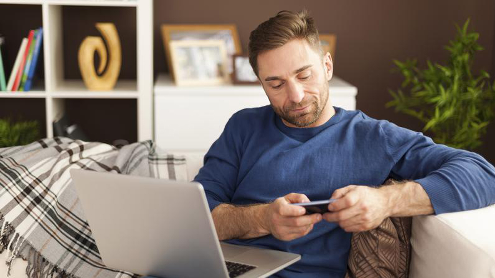 A man sitting on a sofa looking at his mobile phone with a laptop open in front of him.