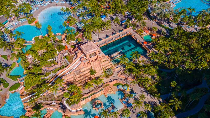 Aerial view of a large resort complex with multiple swimming pools, slides, a pyramid-shaped structure, and surrounding palm trees.