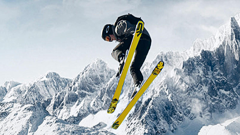 A skier in mid-air performs a trick against a snowy mountain backdrop, holding yellow skis with a clear sky in the background.