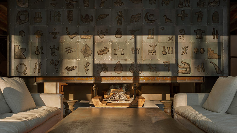 A cozy living room with two beige armchairs and a coffee table facing a modern fireplace. The wall behind features metal art with various abstract shapes and symbols.