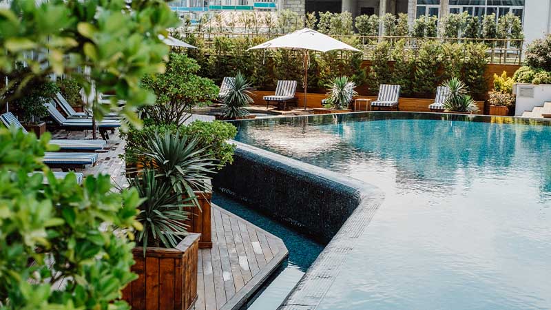 A luxurious outdoor swimming pool with blue water is surrounded by lounge chairs, umbrellas, and greenery, creating a serene atmosphere.